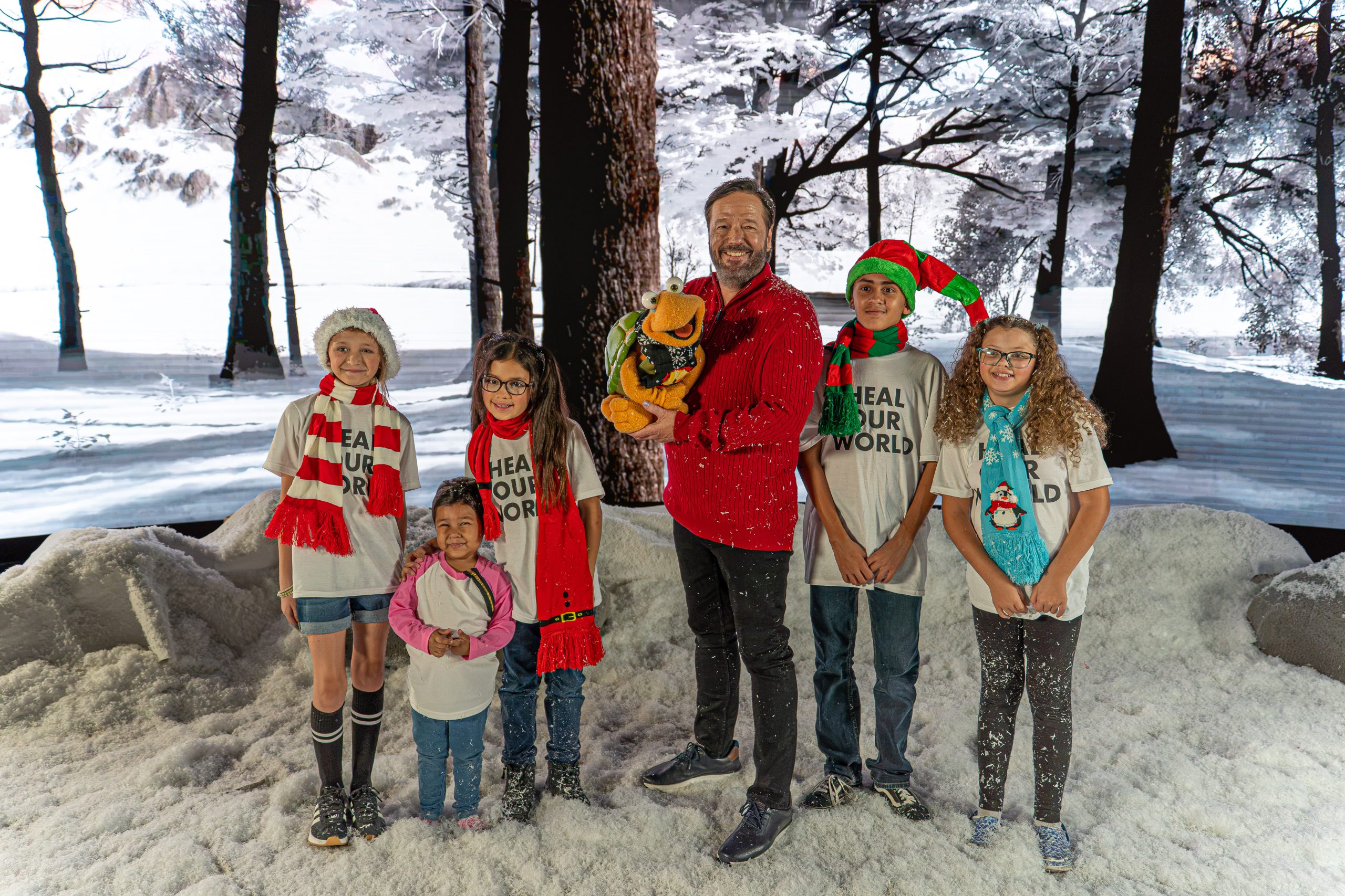 Terry Fator, holding a turtle puppet, stands with five kids from Cure 4 The Kids Foundation on a snowy forest set