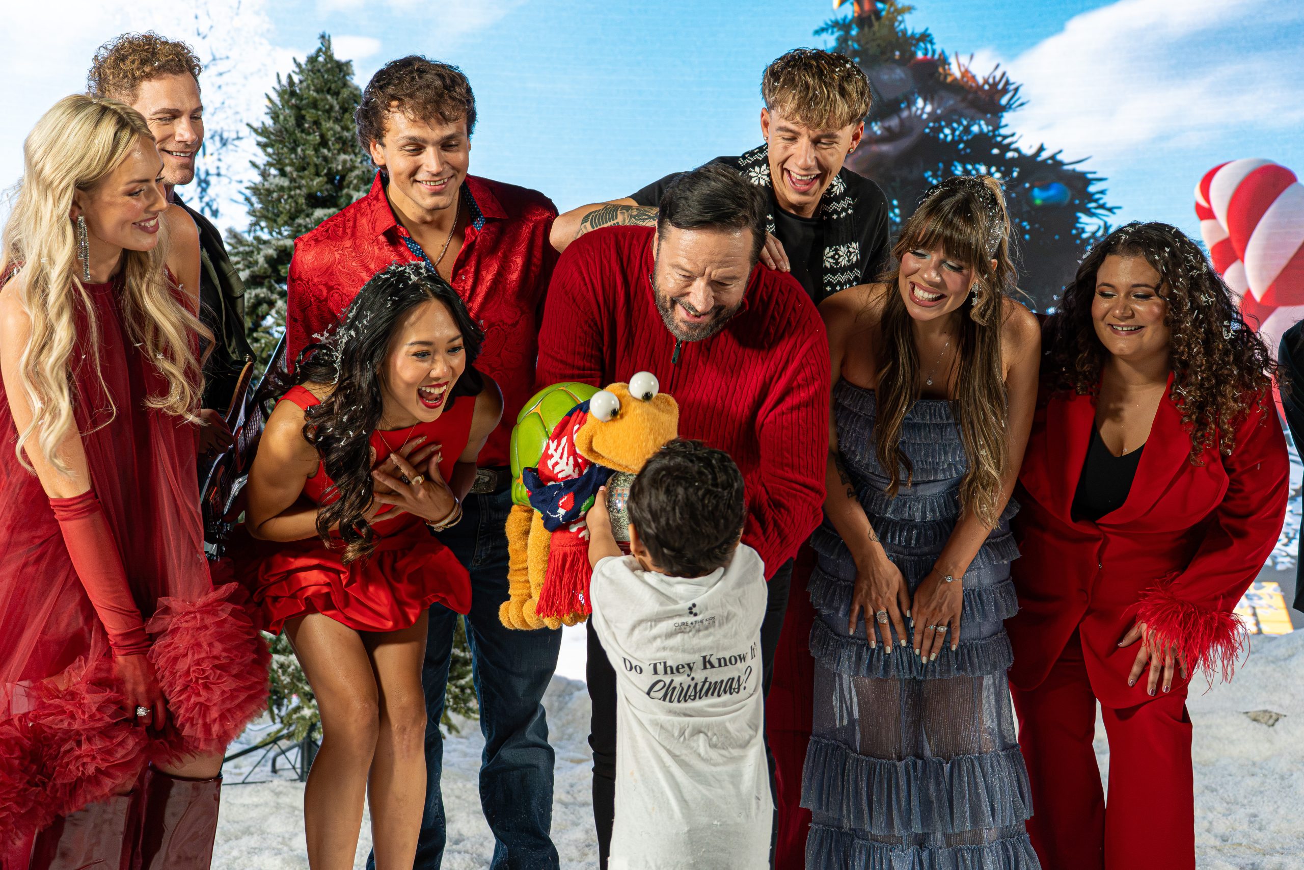 Artists from Do They Know It's Christmas? laugh with a child in a Cure 4 The Kids Foundation shirt and a turtle puppet on a snowy holiday set